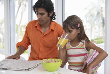 Father reading newspaper sitting with daughter (7-9) having breakfast