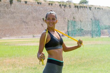 Female water sprinkled wet athlete woman in sportswear measuring her waistline, hips and chest after a workout at an outdoor field stadium. Healthy lifestyle sports and fitness concept.