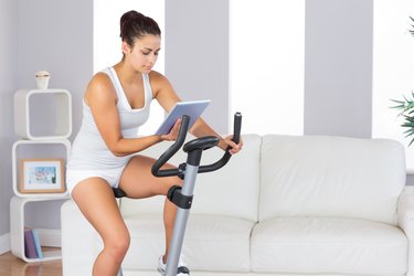 Concentrated slender woman training on an exercise bike while using her tablet