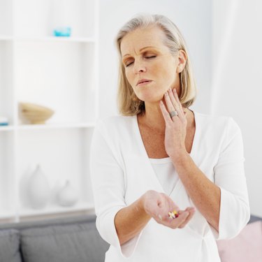 Front view of mature woman holding pills and rubbing her neck with pain
