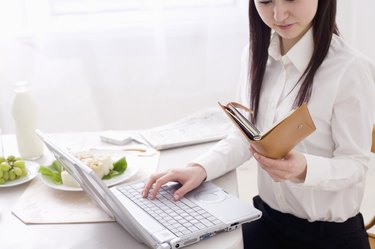 Young woman using laptop at dining table, looking at personal organizer