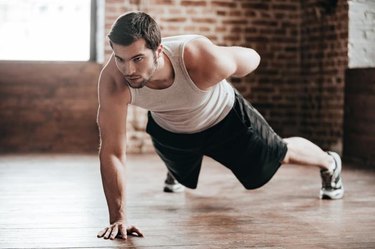 Confident muscled young man wearing sport wear and doing one hand push-up while exercising on the floor in loft interior