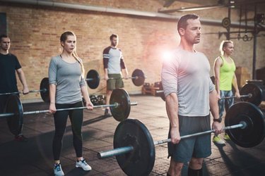 Serious muscular bearded man in shorts and gray tee shirt leading small group of young adults in barbell exercises for fitness training