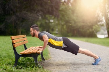 Young man working out doing push-ups on a wooden park bench as he warms up for his daily workout or jog
