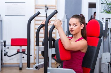 Young, adorable, cute, sexy trainer girl doing her butterfly chest workout exercise on the Pec Deck expressing power and strength in red sportswear