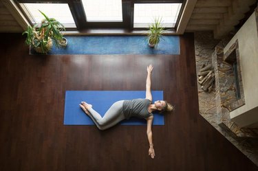 Attractive young woman working out in living room, doing yoga exercise on wooden floor, lying in Belly Twist Pose, Jathara Parivartanasana, resting after practice, full length, top view