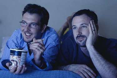 Gay couple lying on bed watching television, one eating ice cream