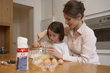 Mother helping daughter (6-8) stir cake mixture in kitchen, smiling