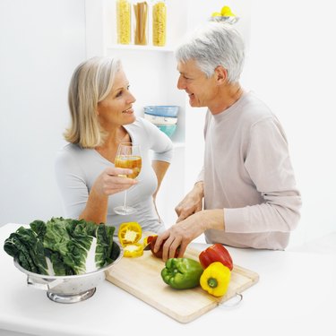 mature man chopping peppers talking to a mature woman who is drinking a glass of white wine