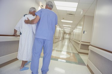 Rear view of male nurse helping senior patient walk in corridor