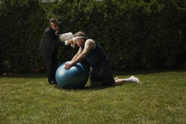 Mature man kneeling on lawn, leaning on exercise ball, butler wiping his forehead
