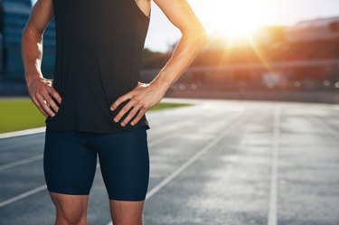 Mid section shot of male athlete standing on race track with his hands on hips on a bright sunlight. Cropped shot of young man runner on athletics running track in stadium.