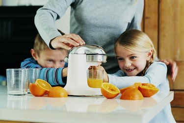 Mother Making Orange Juice in the Kitchen Using a Juicer With Her Son and Daughter Helping Her