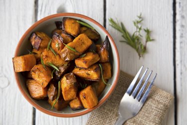 an overhead photo of a bowl of roasted, cut and cubed sweet potatoes with herbs next to a fork on a wooden table