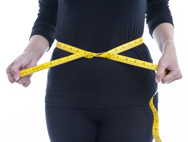 A woman wearing all black wraps a yellow measuring tape around her belly.