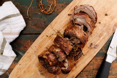 Slices of lamb flaps on a wooden cutting board with a chef's knife and towel