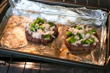 hamburger patties topped with onions and peppers in the oven on foil-lined pan