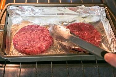 close up of hand holding tongs to flip hamburger patties in oven on foiled-lined pan