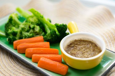 Yellow ramekin of sweet mustard sauce on a green plate of steamed carrots and broccoli.