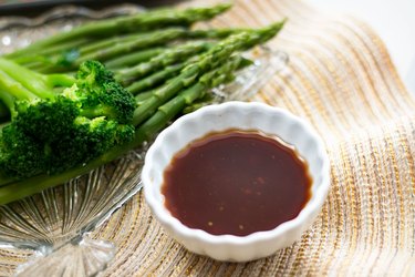 Small bowl of savory brown vinegar sauce served alongside a plate of steamed broccoli and asparagus.