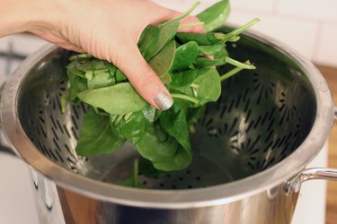 hand placing raw fresh spinach into steaming basket over boiling water