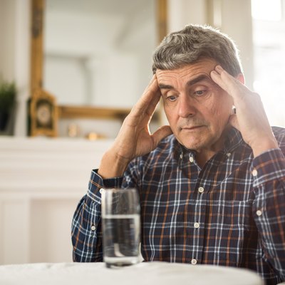An older man sitting at a table with a glass of water, with his hands on his temples because he is feeling dizzy