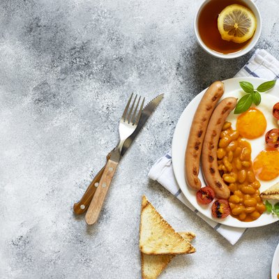 English breakfast with fried sausages, beans, mushrooms, fried eggs, grilled cherry tomatoes. Served with a cup of tea with lemon, bread toast and butter. Gray background, top view, copyspace