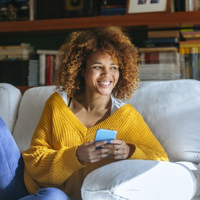 Happy young woman with cell phone sitting on sofa at home