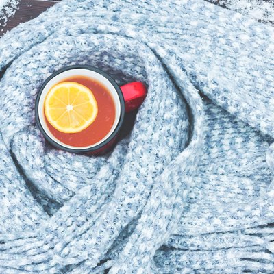 Red enameled cup of hot tea with lemon wrapped in a knitted scarf on a snowy wooden table