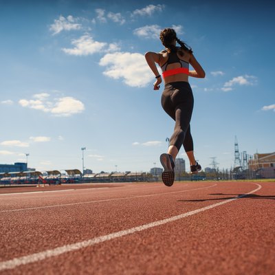 From below back view of female teen athlete in sportswear running on track at stadium on summer day