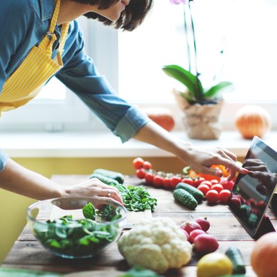 A woman in a yellow apron cooking with vegetables and looking at a recipe on her tablet