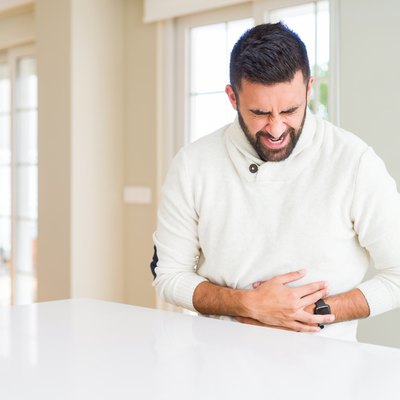 Handsome hispanic man wearing casual white sweater at home with hand on stomach because nausea, painful disease feeling unwell. Ache concept.
