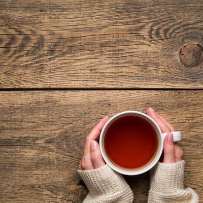 Female hands holding a mug of hot black tea. Cold winter white warm clothes. Top view. Aged rustic wooden background