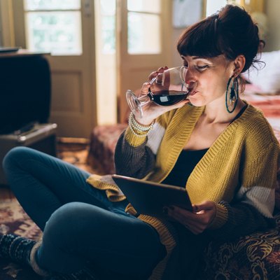 A happy woman drinking a glass of wine at home while using her tablet