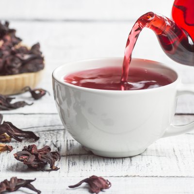 White cup of healthy hibiscus tea pouring from the teapot with dried hibiscus flowers on white wooden background, winter hot drink concept for cold and flu