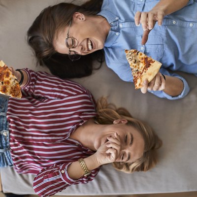 Two laughing young women lying down eating pizza together