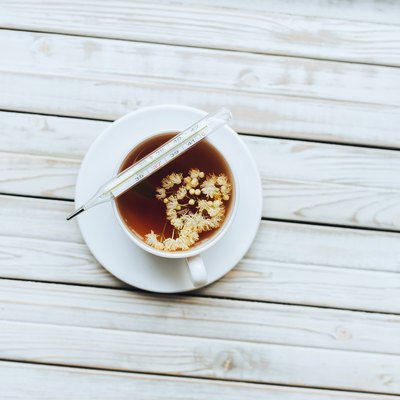 A mercury glass thermometer lies on a white cup with a linden, fragrant healing tea made from dried linden flowers on a grey wooden background.