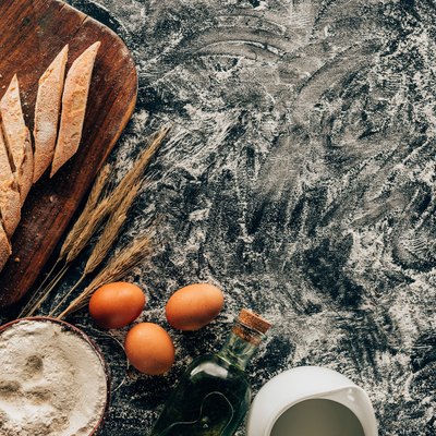 top view of arranged pieces of french baguette, raw eggs, wheat and flour in bowl on dark surface
