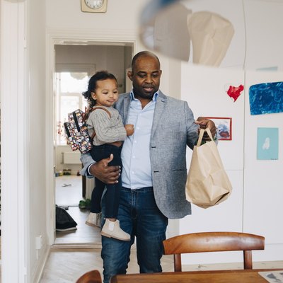 Father carrying daughter with backpack while keeping paper bag on table at home