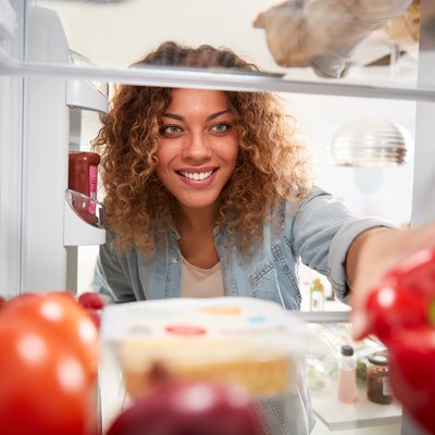 View Looking Out From Inside Of Refrigerator As Woman Opens Door And Unpacks Shopping Bag Of Food