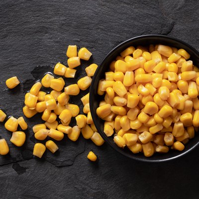 Canned sweet corn in a black ceramic bowl isolated on black slate next to spilled sweet corn. Top view.