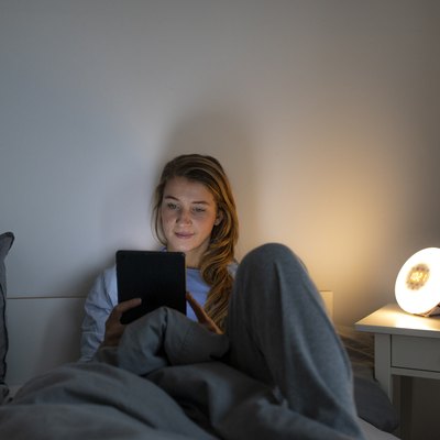 Young woman lying in bed at home at night using tablet