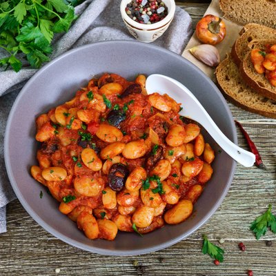 Stewed broad beans in tomato sauce with herbs and spices closeup. Slices of rye bread with beans on the wooden table plant-based recipes