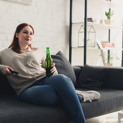 beautiful woman holding bottle of beer and remote control at home