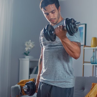 Strong Athletic Fit Man in T-shirt and Shorts is Doing Calf Raise Exercises with Dumbbells at Home in His Spacious and Bright Apartment with Minimalistic Interior.