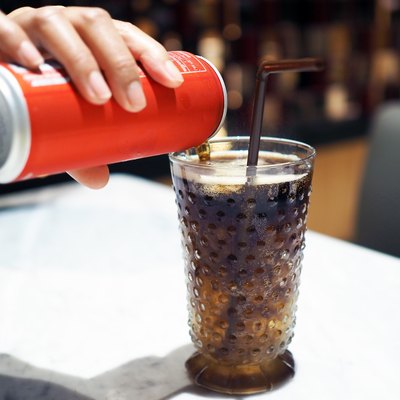 Pouring cola over ice cube in clear glass with straw.