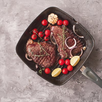 Grilled beef steaks made of marble beef with salt, pepper, thyme, lemon on a grill pan on a gray textured concrete background. The view from the top. Copy space