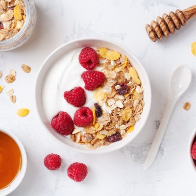 Greek yogurt in bowl with raspberries, honey and muesli on white stone table top view. Healthy diet breakfast.