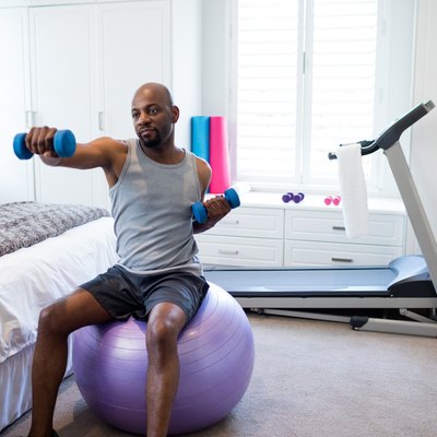 Man exercising with dumbbells on fitness ball in bedroom