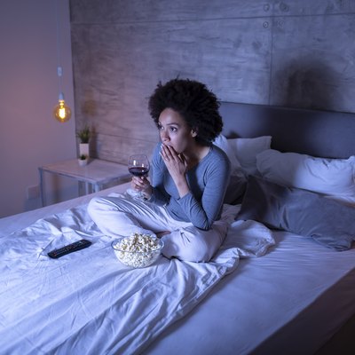 A woman watching a movie on her bed while eating popcorn and drinking a glass of wine